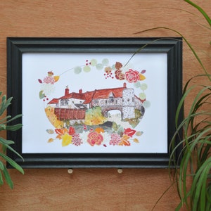 Könnte beinhalten: Ein gerahmtes Aquarellbild eines Gebäudes mit rotem Dach und einem Steinbogen. Das Bild ist von einem Kranz aus Blumen und Blättern in Rot-, Orange-, Gelb- und Grüntönen umgeben.