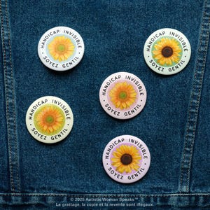 K&ouml;nnte beinhalten: F&uuml;nf runde Buttons mit einem Sonnenblumenmuster und den Worten "HANDICAP INVISIBLE SOYEZ GENTIL" in einem kreisf&ouml;rmigen Muster. Die Buttons sind wei&szlig;, hellblau und rosa, vor einem Jeanshintergrund.
