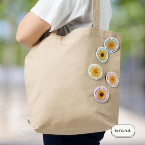 K&ouml;nnte beinhalten: Eine beige Tragetasche mit mehreren runden Anstecknadeln. Die Anstecknadeln zeigen ein Sonnenblumen-Design mit den Worten "HANDICAP INVISIBLE SOYEZ GENTIL" in einem kreisf&ouml;rmigen Muster. Das Wort "Grand" ist ebenfalls sichtbar.