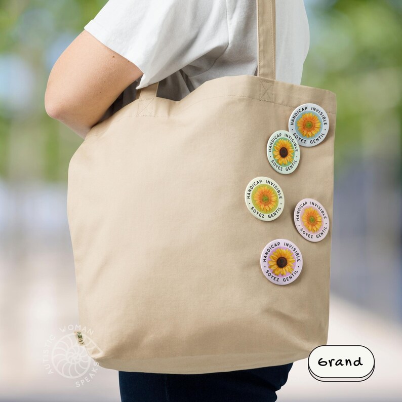 K&ouml;nnte beinhalten: Eine beige Tragetasche mit mehreren runden Anstecknadeln. Die Anstecknadeln zeigen ein Sonnenblumen-Design mit den Worten "HANDICAP INVISIBLE SOYEZ GENTIL" in einem kreisf&ouml;rmigen Muster. Das Wort "Grand" ist ebenfalls sichtbar.