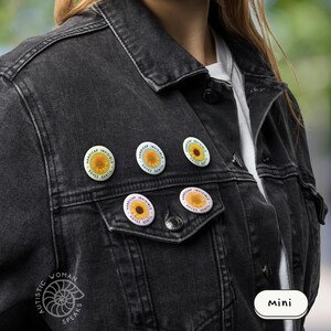 K&ouml;nnte beinhalten: Eine schwarze Jeansjacke, verziert mit mehreren runden Anstecknadeln. Jede Anstecknadel zeigt ein Sonnenblumen-Design mit den Worten "Handicap Invisible Soyez Gentil" in kreisf&ouml;rmiger Anordnung. Die Jacke hat ein klassisches Design mit Taschen und Kn&ouml;pfen.