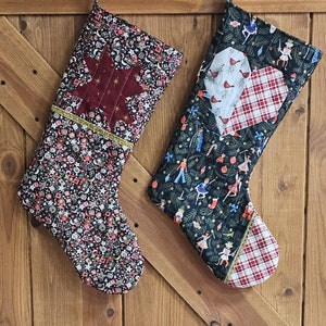 May include: Two quilted Christmas stockings hanging on a wooden door. One stocking features a red star with gold glitter and the other has a red and white plaid heart with a bird design.