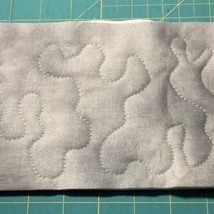 May include: A rectangular piece of gray fabric with a mottled pattern, featuring a quilted design of flowing, organic shapes in white thread. The fabric is layered over a white backing, creating a textured effect.