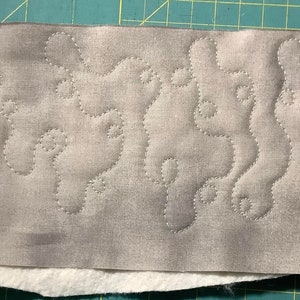 May include: A rectangular piece of fabric with a gray, patterned top layer and a white bottom layer. The gray fabric features a stitched, flowing design in a lighter gray thread. The layers are stacked on top of each other.