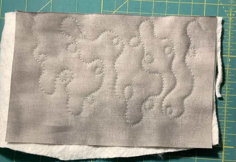 May include: A rectangular piece of fabric with a gray, patterned top layer and a white bottom layer. The gray fabric features a stitched, flowing design in a lighter gray thread. The layers are stacked on top of each other.