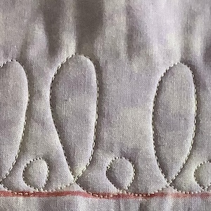 May include: Close-up of a quilted fabric with a repeating pattern of teardrop shapes. The shapes are outlined with white stitching on a light purple background. A red line runs horizontally near the bottom of the image, adding a contrasting element to the design.