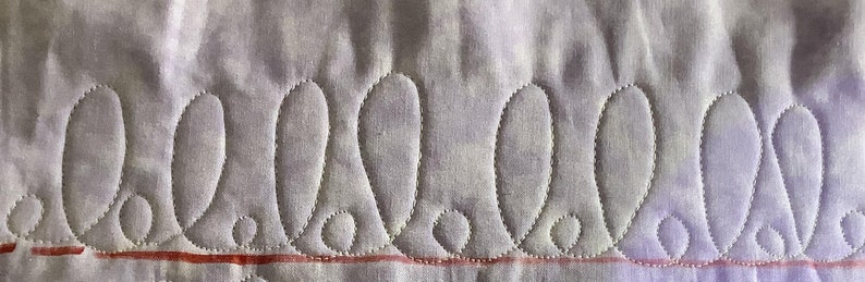 May include: Close-up of a quilted fabric with a repeating pattern of teardrop shapes. The shapes are outlined with white stitching on a light purple background. A red line runs horizontally near the bottom of the image, adding a contrasting element to the design.