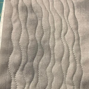 May include: Close-up of a gray fabric with a wavy, quilted pattern. The stitching is white, creating a contrasting design. The fabric appears to be a light, neutral color, suitable for various sewing projects.