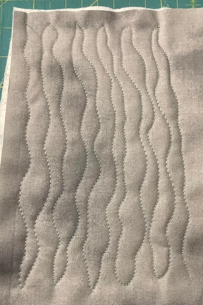 May include: Close-up of a gray fabric with a wavy, quilted pattern. The stitching is white, creating a contrasting design. The fabric appears to be a light, neutral color, suitable for various sewing projects.