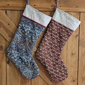May include: Two Christmas stockings with floral patterns hang against a wooden background. One stocking has a navy blue background with light blue and peach floral designs. The other stocking has a red background with white and brown floral designs.