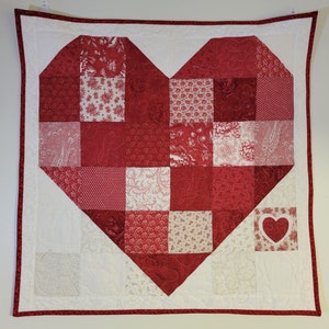 May include: A red and white patchwork quilt with a large heart shape in the center. The heart is made of smaller squares of fabric in various red and white patterns. The quilt is edged with a red border.