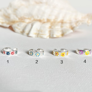 Könnte beinhalten: Vier silberne Zehenringe mit farbenfrohen Blumenmustern. Jeder Ring hat ein anderes Design: rot, blau und rosa; blau und gelb; gelb; und lila und gelb. Die Ringe werden auf weißem Hintergrund gezeigt.