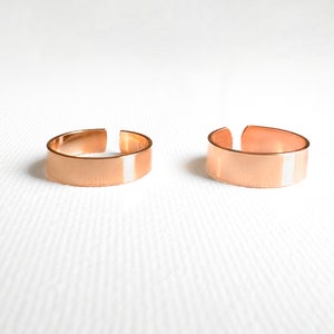 May include: Two copper-coloured adjustable rings. Each ring has a flat band design with an open front. The rings are displayed on a white surface. The rings are likely made of copper or a copper alloy.
