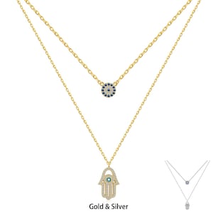 Könnte beinhalten: Eine mehrreihige Halskette aus Gold und Silber mit einem Hamsa-Hand-Anhänger und einem runden Anhänger mit einem blauen bösen Auge. Die Anhänger sind mit klaren und blauen Kristallen verziert. Die Halskette ist mit "Gold & Silber" beschriftet.