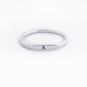 May include: A silver ring with a smooth, wavy design. The ring features the letter "A" engraved on the front. The ring is set against a white background.