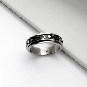 Peut inclure: Une bague tournante en argent avec une bande noire ornée d'un motif soleil, lune et étoiles. L'intérieur de la bande est gravé avec "A♥M".