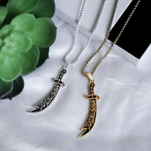 Könnte beinhalten: Zwei silberne und goldene Halsketten mit einem Schwertpendant. Der Schwertpendant ist schwarz mit arabischer Schrift.