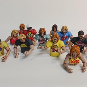 May include: A collection of miniature figurines, each depicting a torso with arms, wearing various band t-shirts. The figurines have different hair colors and styles. The shirts feature logos from bands like AC/DC, The Rolling Stones, and Harley-Davidson.