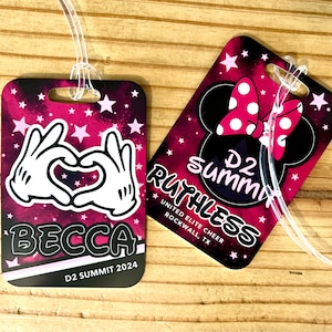 May include: Two rectangular luggage tags with clear plastic loops. One tag features a heart made of white gloved hands, the name "BECCA", and "D2 SUMMIT 2024". The other tag has a Minnie Mouse bow and the text "D2 Summit RUTHLESS". Both tags have a pink and black background.