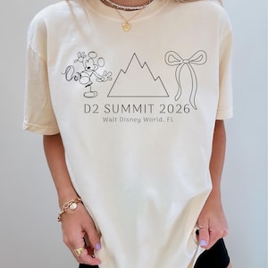 May include: Cream-colored t-shirt with a black line drawing of Mickey Mouse, a mountain range, and a bow. The text "D2 SUMMIT 2026 Walt Disney World, FL" is printed below the graphics. The shirt has short sleeves and a crew neck.