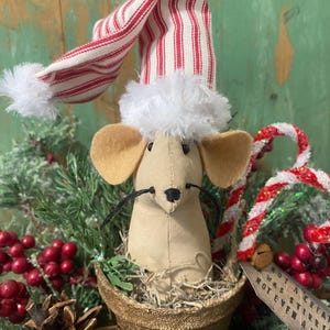 May include: A whimsical Christmas decoration featuring a tan mouse wearing a red and white striped hat with a white fluffy trim. The mouse sits in a small brown pot, surrounded by greenery, red berries, and pinecones. A candy cane and a small tag are also visible.