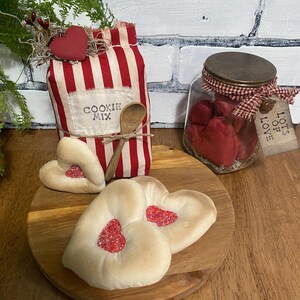 May include: A rustic display featuring a red and white striped bag labeled "COOKIE MIX" with a wooden spoon, heart-shaped cookies, and a jar filled with red hearts. A wooden heart decorates the bag.