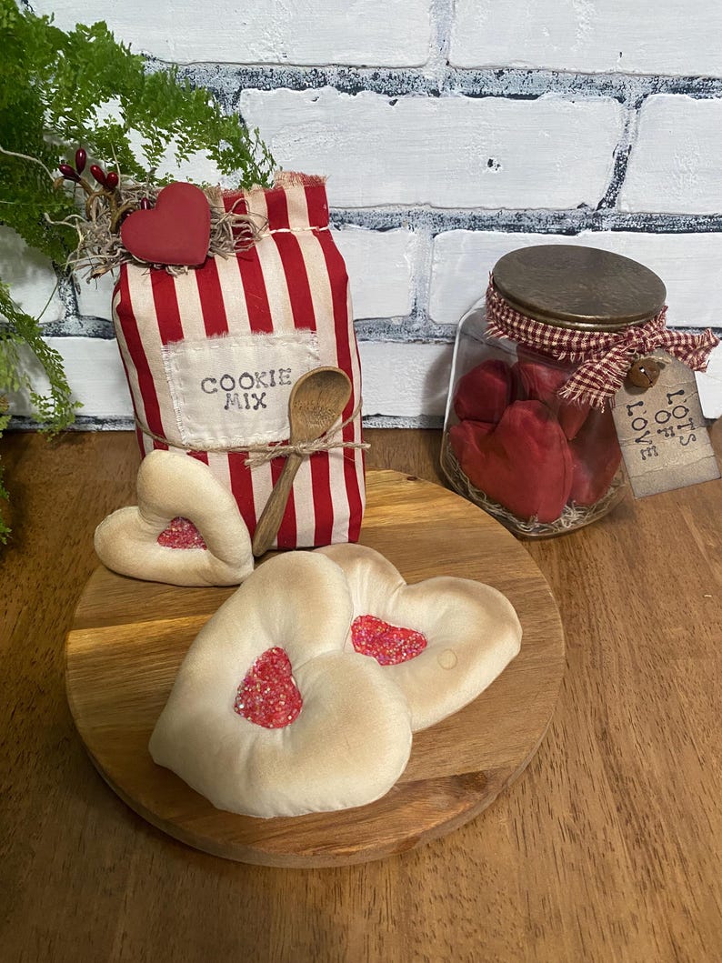 May include: A rustic display featuring a red and white striped bag labeled "COOKIE MIX" with a wooden spoon, heart-shaped cookies, and a jar filled with red hearts. A wooden heart decorates the bag.
