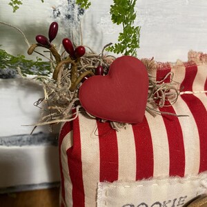 May include: A decorative cookie mix package with a red heart and red and white striped fabric. The package is adorned with sprigs of greenery and berries. The text "COOKIE MIX" is visible on a label.