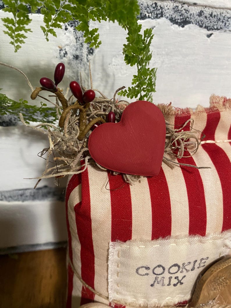 May include: A decorative cookie mix package with a red heart and red and white striped fabric. The package is adorned with sprigs of greenery and berries. The text "COOKIE MIX" is visible on a label.