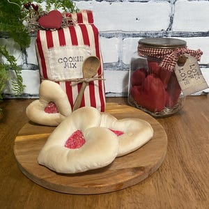May include: A rustic Valentine's Day display featuring a red and white striped cookie mix bag, a wooden spoon, heart-shaped cookies, and a jar filled with red heart-shaped candles. A wooden cutting board and a tag that says "Lots of Love" are also included.