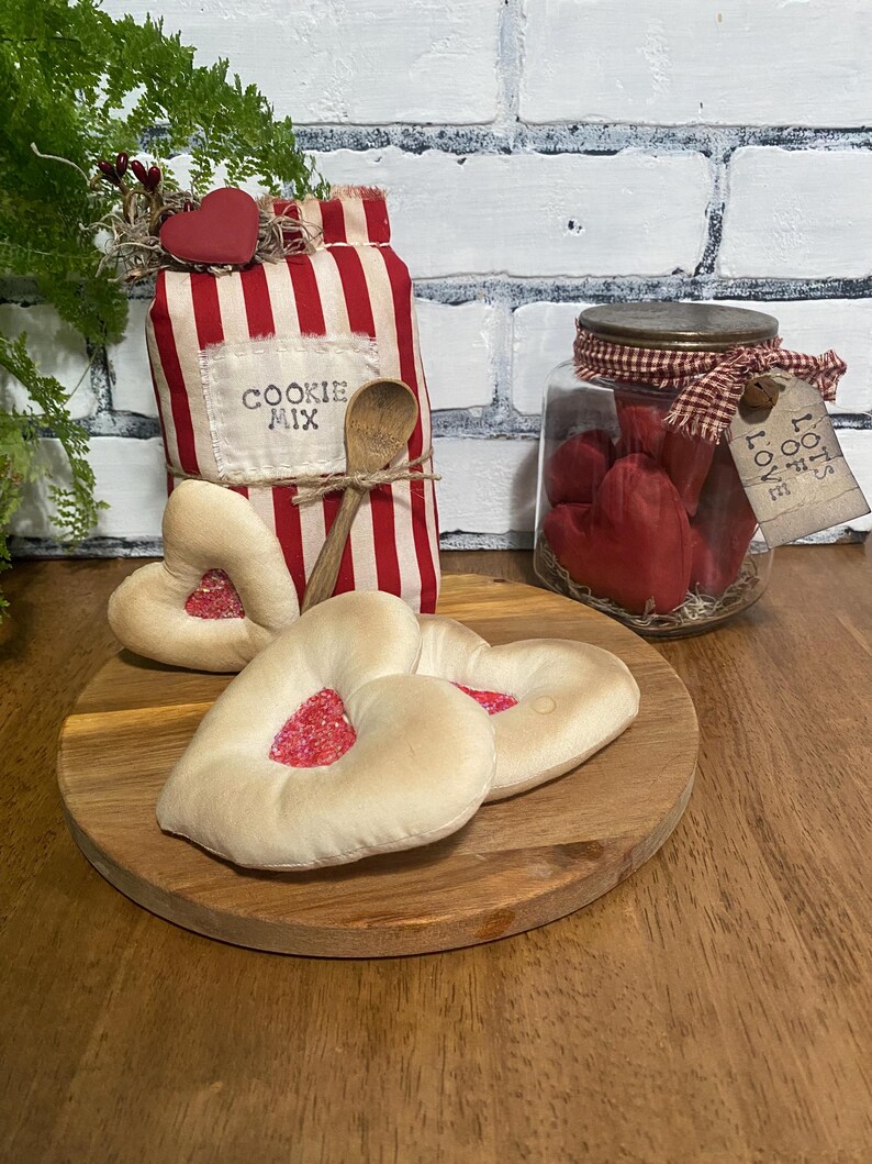 May include: A rustic Valentine's Day display featuring a red and white striped cookie mix bag, a wooden spoon, heart-shaped cookies, and a jar filled with red heart-shaped candles. A wooden cutting board and a tag that says "Lots of Love" are also included.