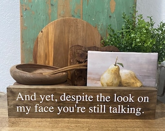 May include: Wooden sign with the text "And yet, despite the look on my face you're still talking."