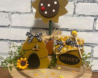 May include: A wooden sunflower, beehive, and honey jar set on a wooden cake stand. The sunflower has a brown center with white dots and a ladybug. The beehive has a bee and the honey jar says "HONEY".
