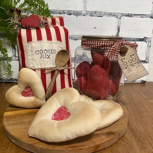 May include: A rustic Valentine's Day display featuring a jar of red hearts, heart-shaped cookies, and a cookie mix bag with a wooden spoon. The items are arranged on a wooden tray, with a brick background.