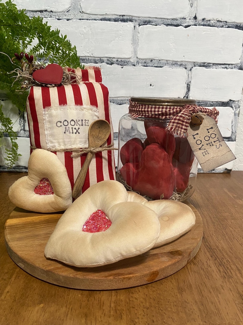 May include: A rustic Valentine's Day display featuring a jar of red hearts, heart-shaped cookies, and a cookie mix bag with a wooden spoon. The items are arranged on a wooden tray, with a brick background.