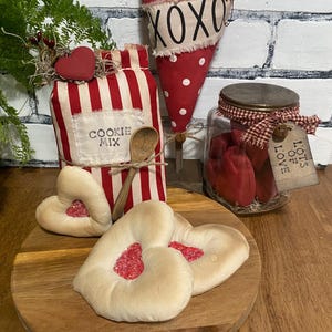 May include: A wooden tray holds heart-shaped cookies with red filling, a jar of red hearts, and a red and white striped container labeled "COOKIE MIX". A red heart with white polka dots and "XOXO" text is also present.