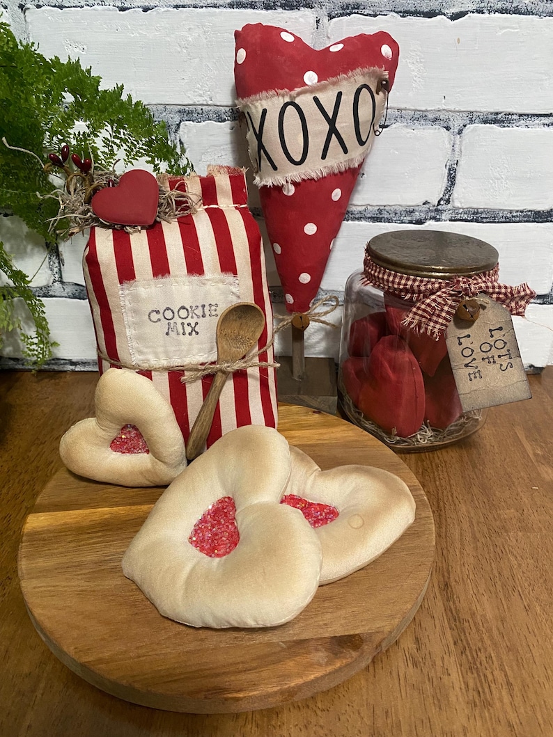 May include: A wooden tray holds heart-shaped cookies with red filling, a jar of red hearts, and a red and white striped container labeled "COOKIE MIX". A red heart with white polka dots and "XOXO" text is also present.