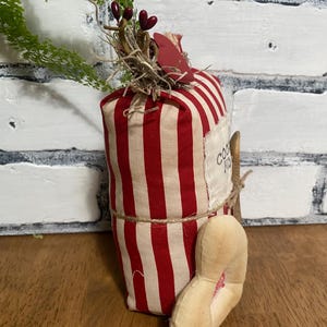 May include: A decorative fabric carrot with red and cream stripes, adorned with greenery, berries, and a small heart-shaped tag. The carrot is tied with twine and has a fabric foot attached. The text "COOKIES" is visible on a label.