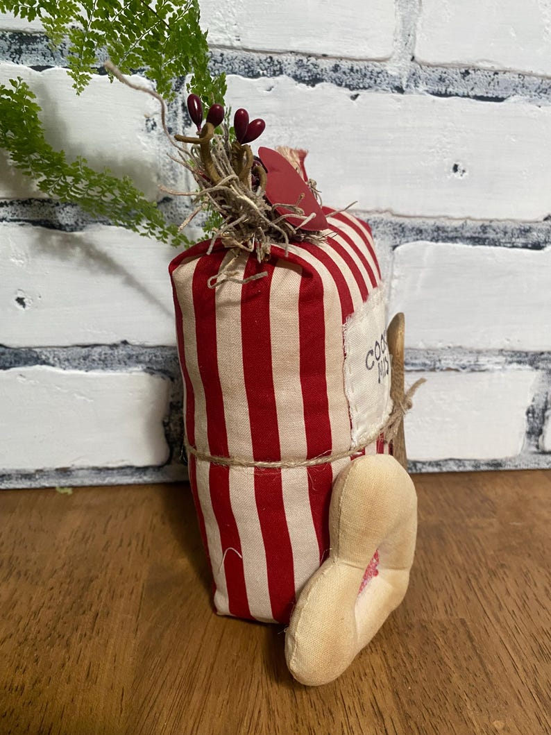 May include: A decorative fabric carrot with red and cream stripes, adorned with greenery, berries, and a small heart-shaped tag. The carrot is tied with twine and has a fabric foot attached. The text "COOKIES" is visible on a label.