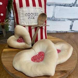 May include: A decorative cookie mix container with red and white stripes, a wooden spoon, and heart-shaped decorations. The container has the words "COOKIE MIX" on a label. The hearts have a red glitter center and sit on a wooden board.