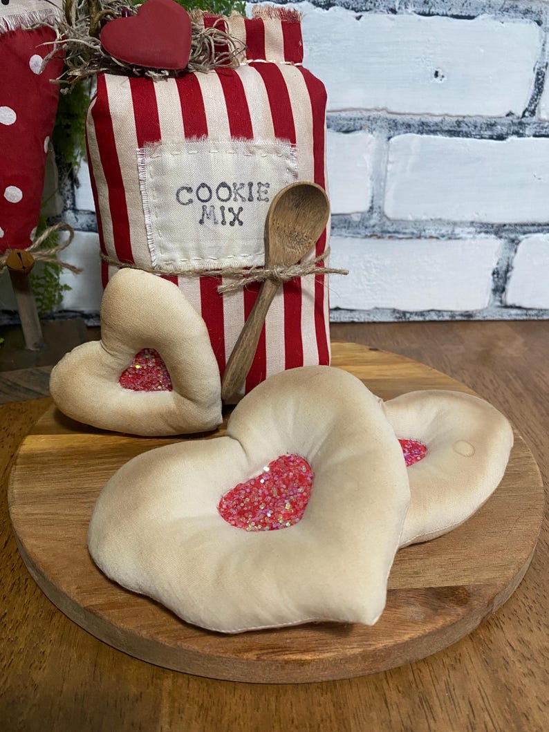 May include: A decorative cookie mix container with red and white stripes, a wooden spoon, and heart-shaped decorations. The container has the words "COOKIE MIX" on a label. The hearts have a red glitter center and sit on a wooden board.