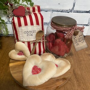 May include: A rustic display featuring a red and white striped cookie mix container, a glass jar filled with red hearts, and heart-shaped cookies on a wooden plate. A wooden spoon and a tag that says "Lots of Love" are also included.