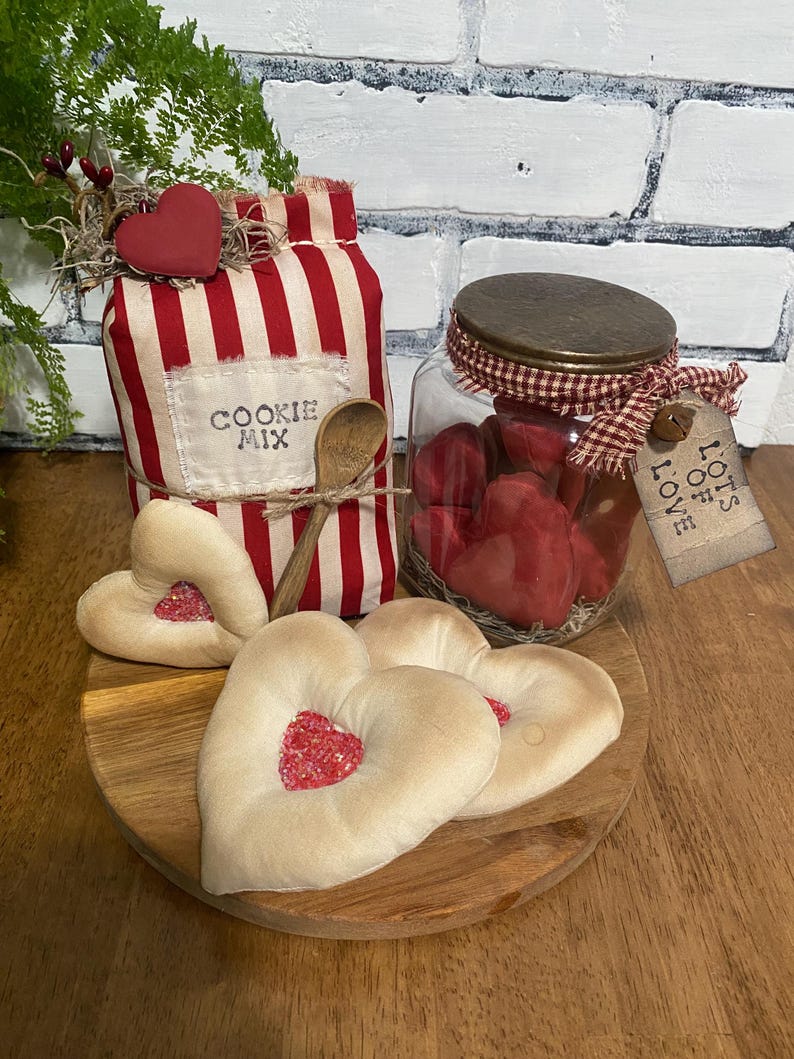 May include: A rustic display featuring a red and white striped cookie mix container, a glass jar filled with red hearts, and heart-shaped cookies on a wooden plate. A wooden spoon and a tag that says "Lots of Love" are also included.