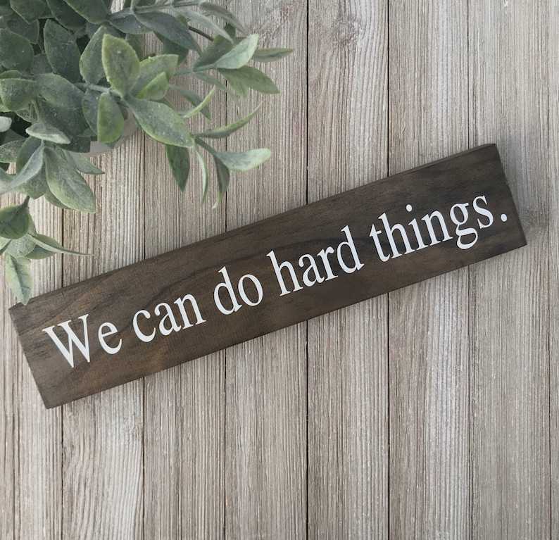 May include: A wooden sign with the text "We can do hard things." painted in white on a dark brown background.