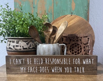May include: Wooden sign with the text "I can't be held responsible for what my face does when you talk". The sign is sitting on a wooden table with a potted plant, wooden spoons, and a ceramic pitcher in the background.
