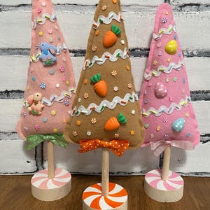 May include: Three felt Easter trees decorated with pastel colors, sprinkles, and Easter-themed embellishments. The trees are on wooden stands with candy cane-style bases.