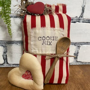 May include: A decorative cookie mix package with red and white stripes, a wooden spoon, and a heart-shaped ornament. The package has the words "COOKIE MIX" printed on it. A second heart-shaped ornament with a glitter-filled center is also present.