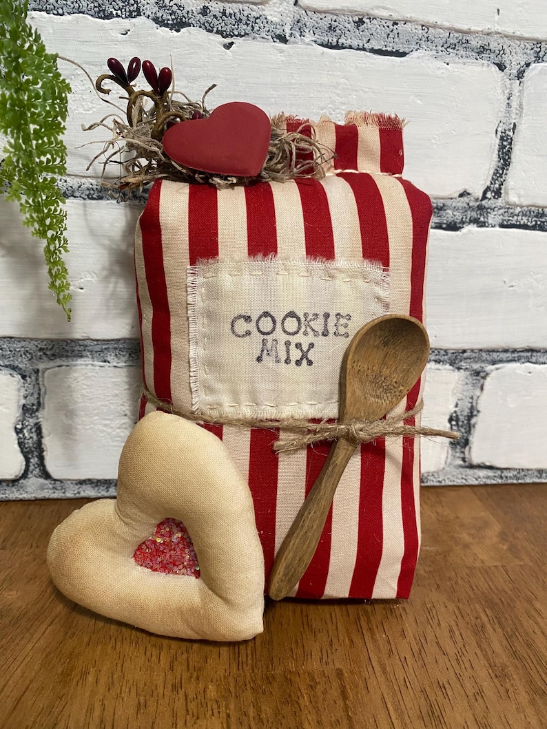 May include: A decorative cookie mix package with red and white stripes, a wooden spoon, and a heart-shaped ornament. The package has the words "COOKIE MIX" printed on it. A second heart-shaped ornament with a glitter-filled center is also present.