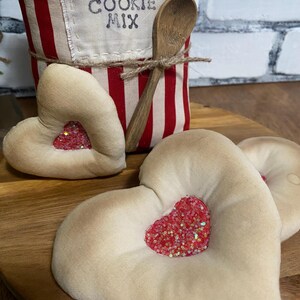 May include: A wooden serving tray holds three heart-shaped fabric decorations with glittery red heart centers. A red and white striped container labeled "COOKIE MIX" and a wooden spoon are in the background.