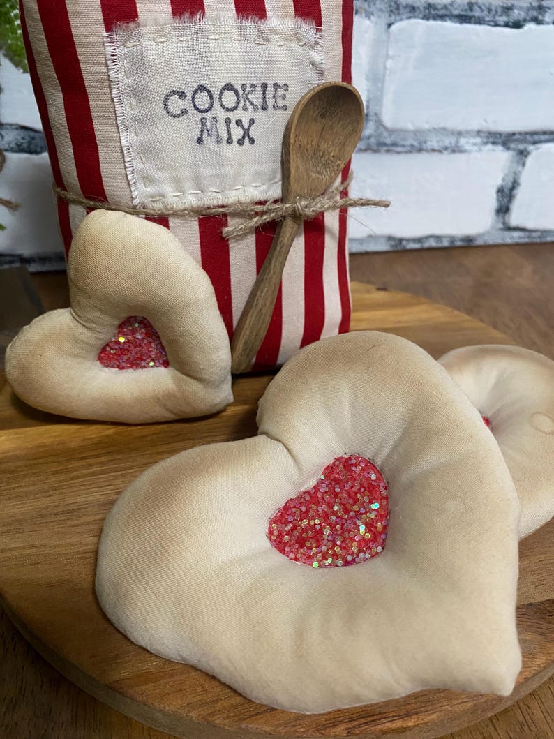 May include: A wooden serving tray holds three heart-shaped fabric decorations with glittery red heart centers. A red and white striped container labeled "COOKIE MIX" and a wooden spoon are in the background.